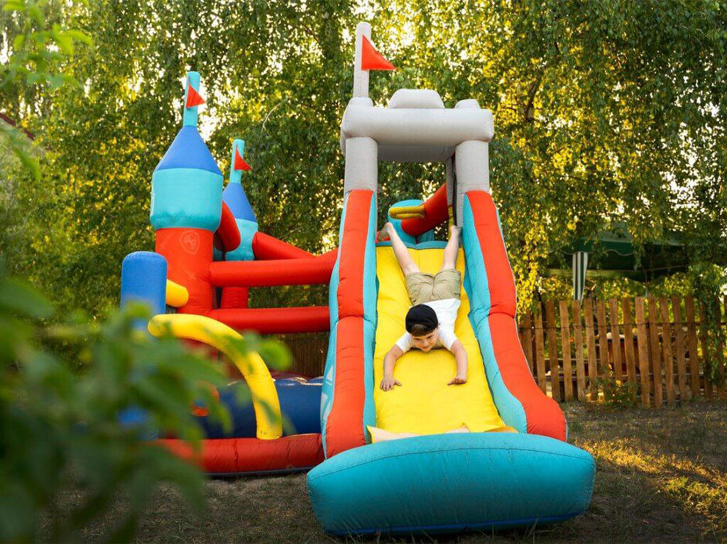A child sliding down a colorful inflatable slide in an outdoor play area, surrounded by greenery.