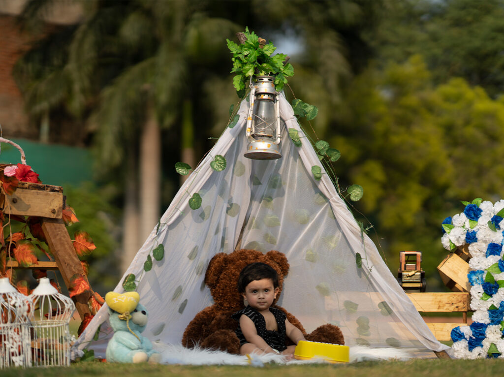 A baby sitting inside a beautifully decorated outdoor teepee with a teddy bear, lantern, and floral accents, organized by Kids Day Events.