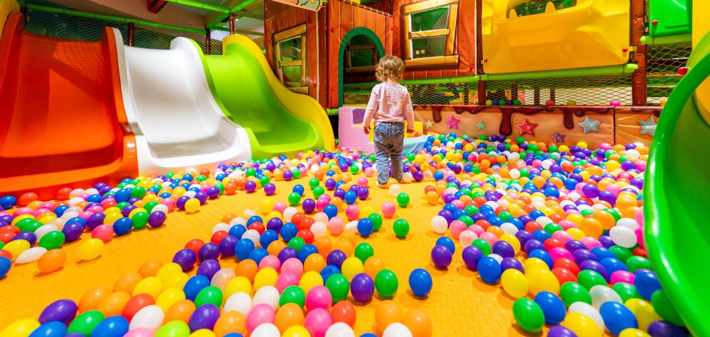 A toddler walking in a colorful indoor ball pit surrounded by slides.