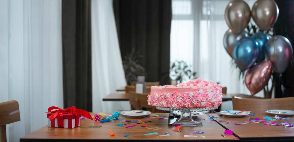 Pink birthday cake on a decorated table with confetti, balloons, and a gift box in a festive party setting.