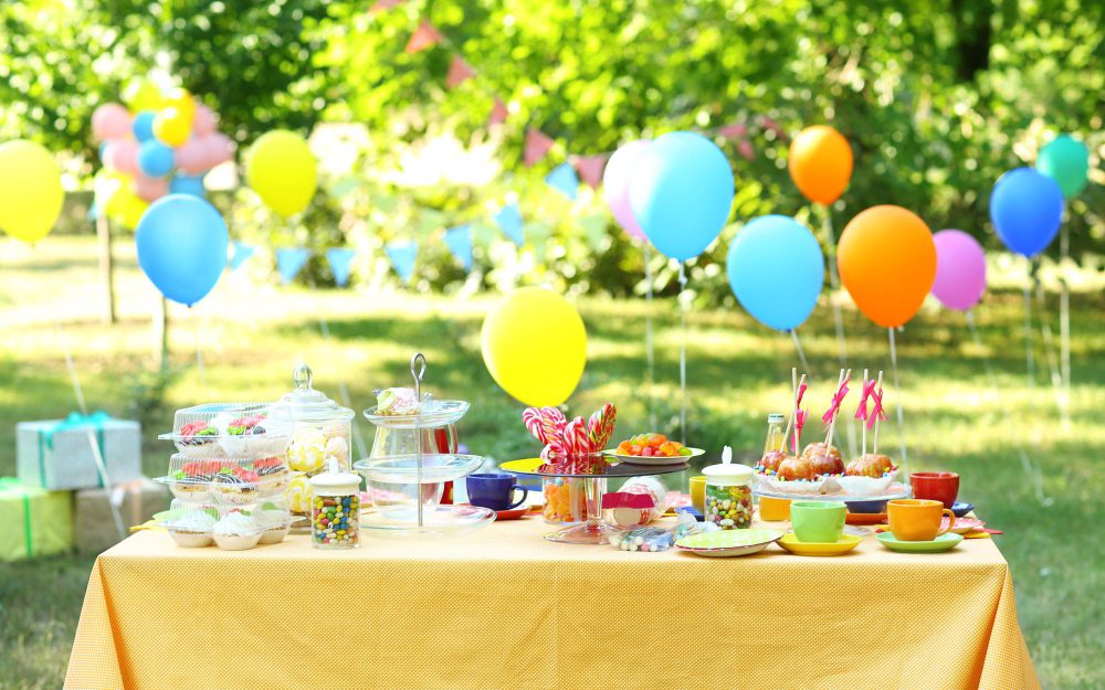 Outdoor birthday party table with colorful balloons, cupcakes, candies, and drinks in a garden setting.