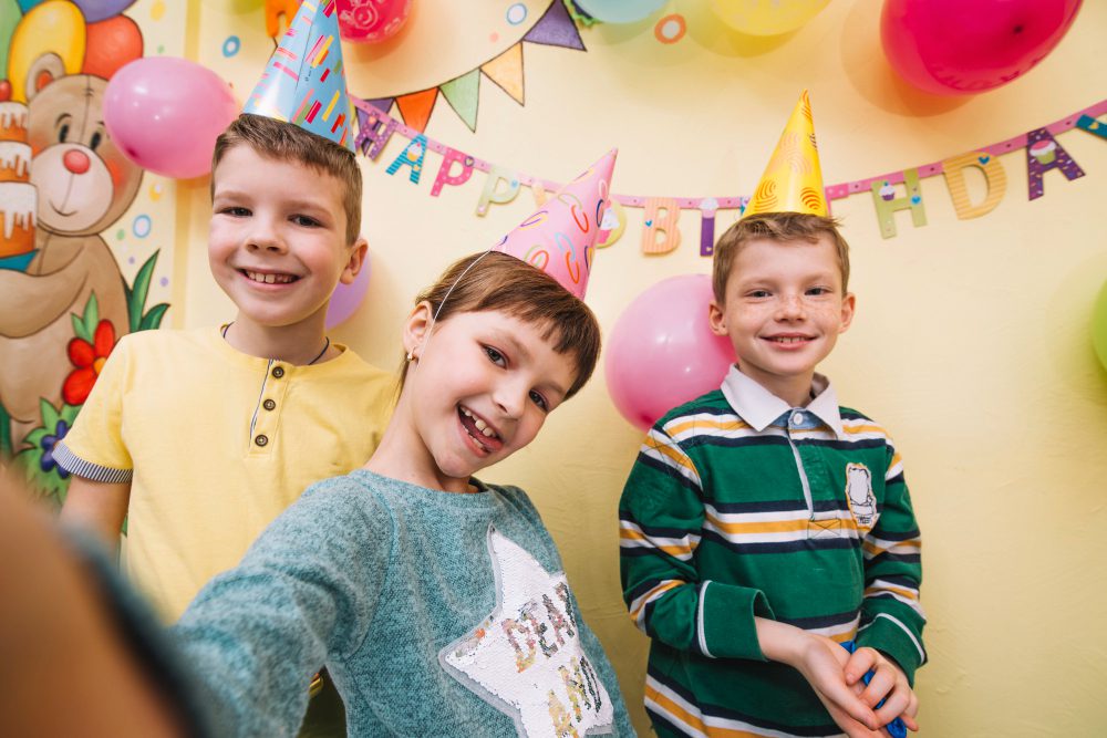 Three happy kids wearing birthday hats and smiling at a colorful birthday party with balloons and decorations